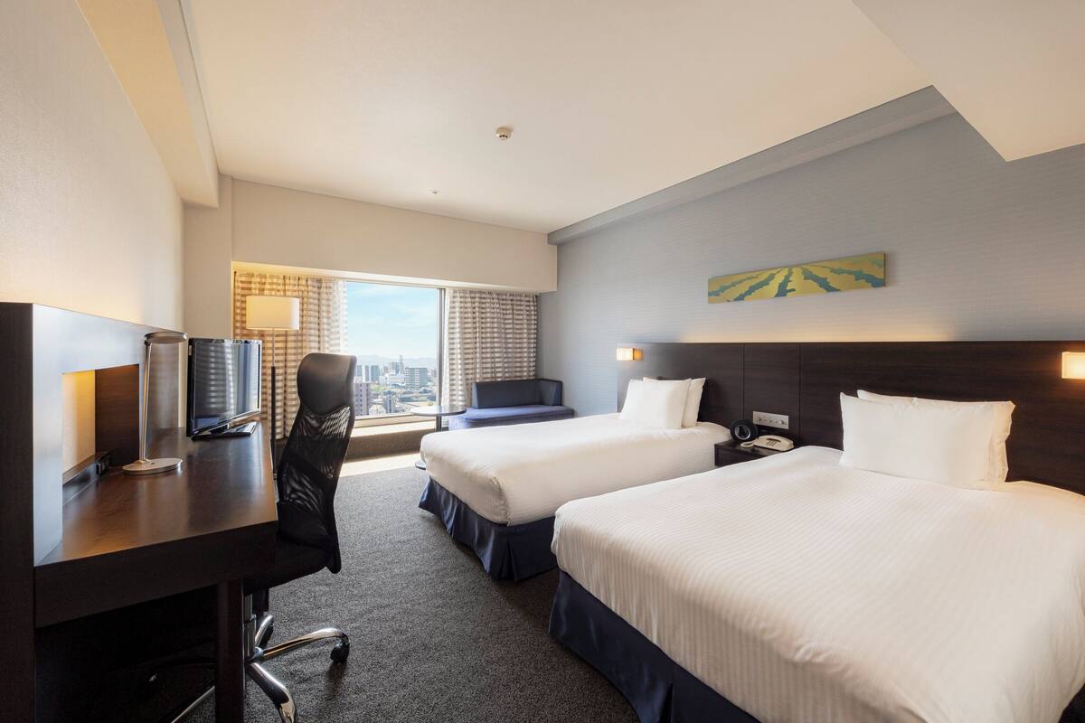 Ein Hotelzimmer mit zwei Betten, einem Schreibtisch, einem Stuhl, einem Fernseher und einem Fenster mit Blick auf die Stadt.