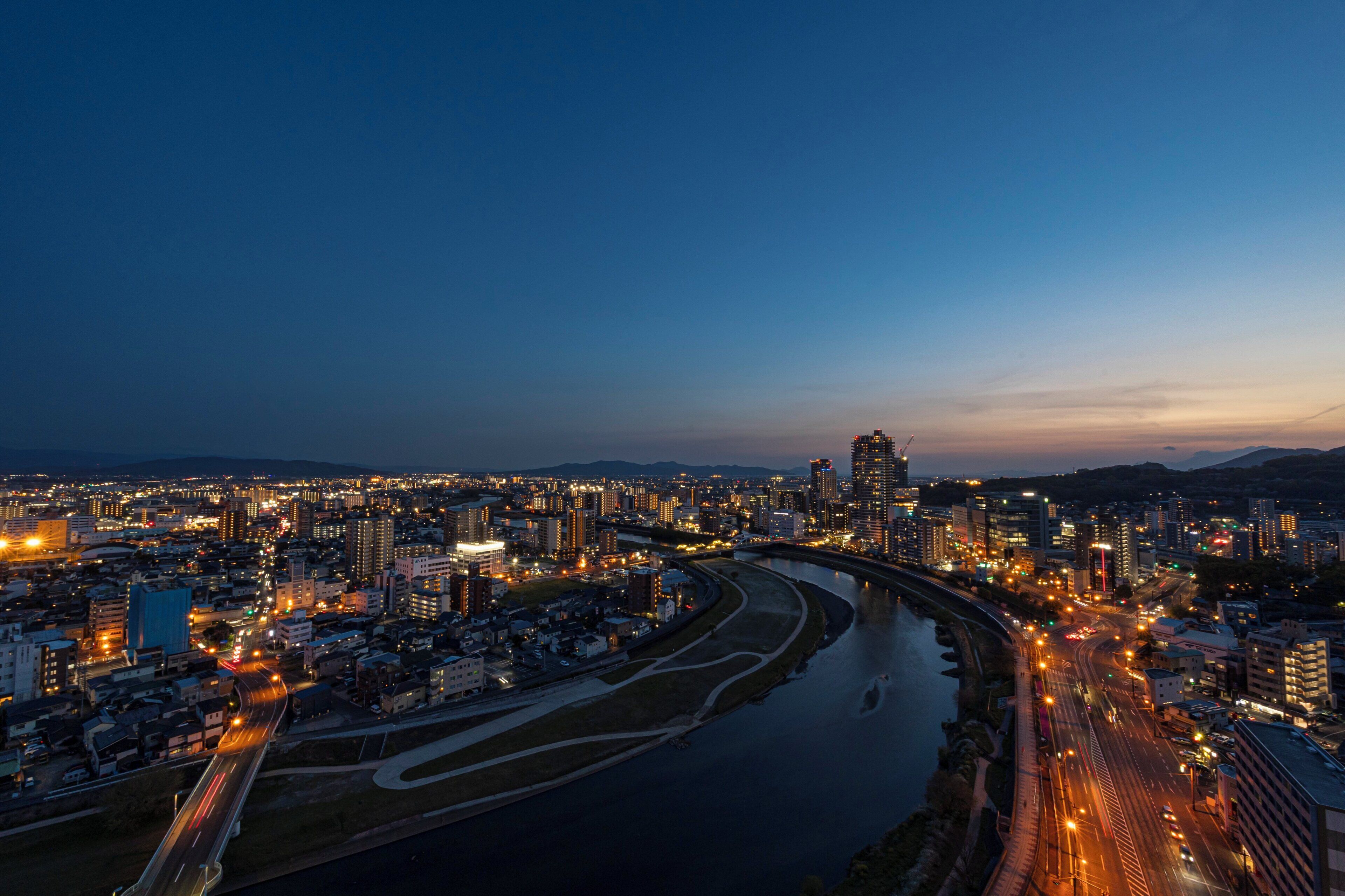A cityscape at dusk with a river running through the center, surrounded by high-rise buildings and illuminated streets.