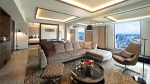 Una habitación de hotel moderna con un amplio sofá modular, una mesa de centro redonda con un florero y copas de vino, un sillón estampado y vistas de la ciudad a través de grandes ventanales.