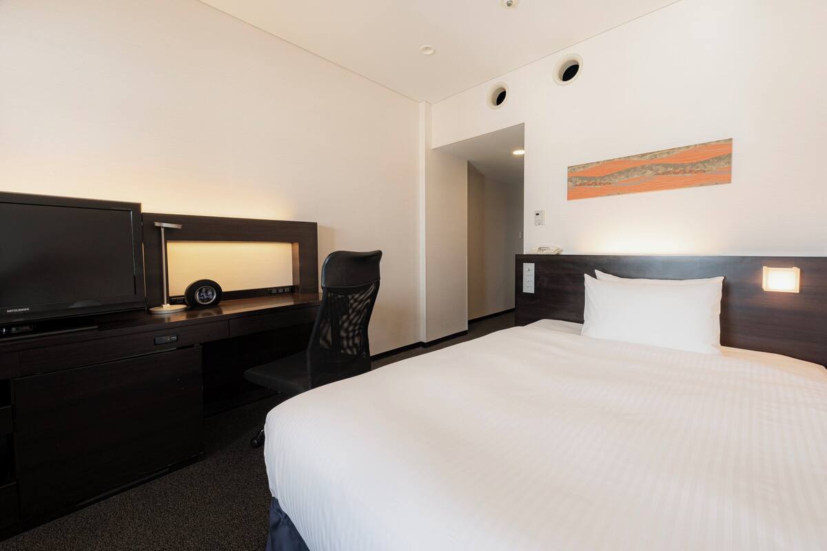 Ein Hotelzimmer mit einem großen Bett, einem Schreibtisch mit Fernseher, einem Sessel und einem Bild an der Wand.