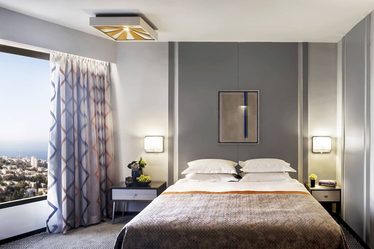 A hotel room with a large bed, two bedside tables, a nightstand, a wall-mounted light, and a view of the city.