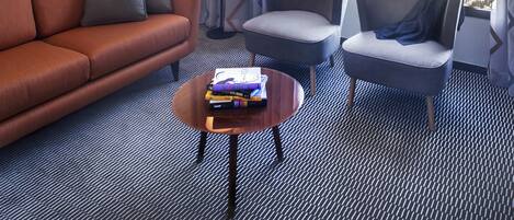 Une chambre d’hôtel moderne dotée d’une grande fenêtre donnant sur la ville, d’un canapé, de deux fauteuils, d’une petite table basse avec des livres et d’un tapis à motifs.