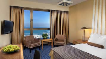 Ein Hotelzimmer mit großem Fenster, zwei Stühlen, einem kleinen Tisch mit Pflanze, einem Fernseher und einem Bett mit gemustertem Überwurf.