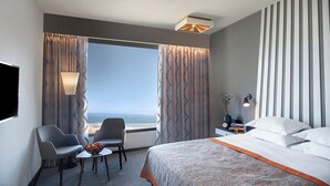 Une chambre d’hôtel avec un lit, deux chaises, une petite table sur laquelle se trouve un saladier de fruits, une table de chevet avec des livres et un vase, et une grande fenêtre avec des rideaux.