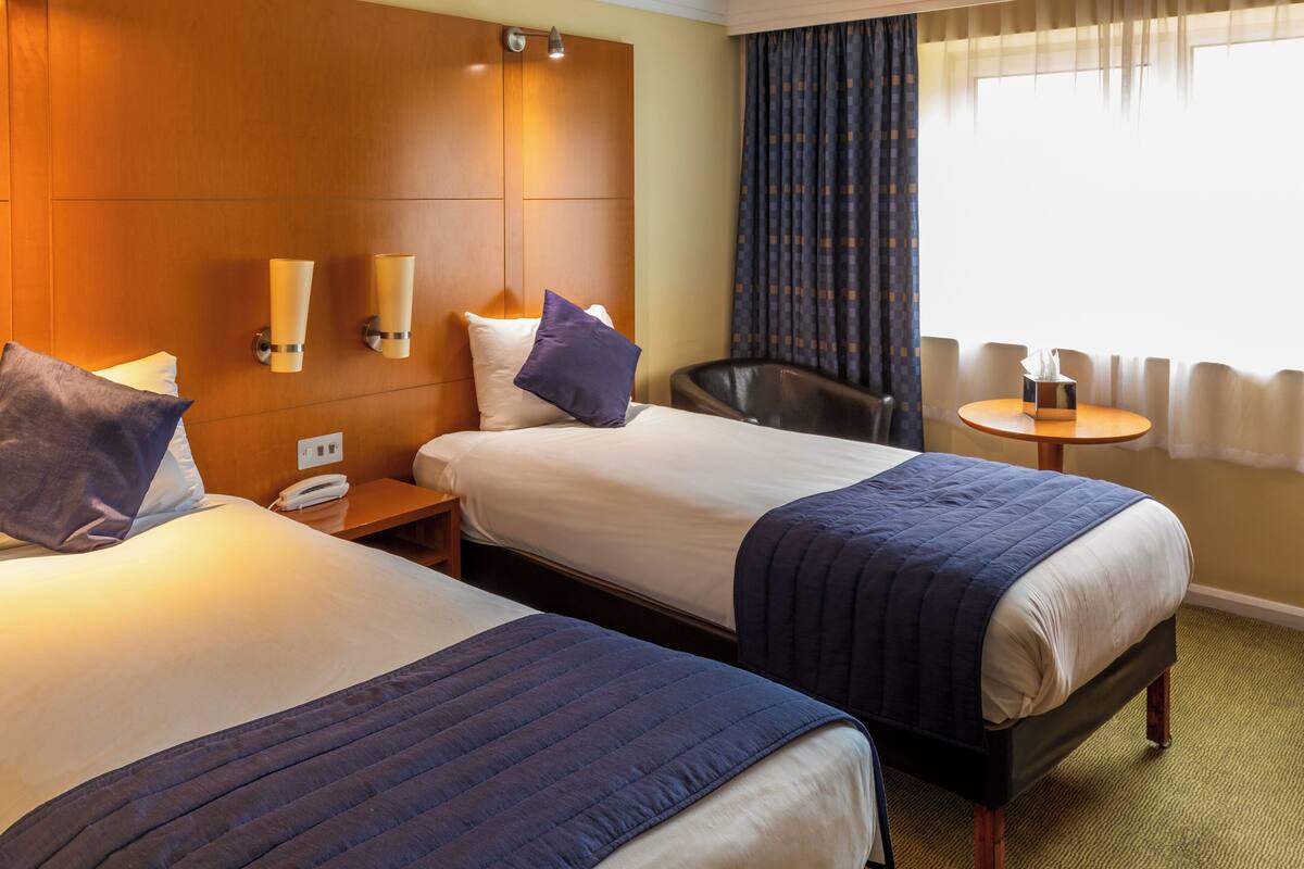 A hotel room with two beds, a wooden headboard, bedside lamps, a chair, a small table, and a window with curtains.