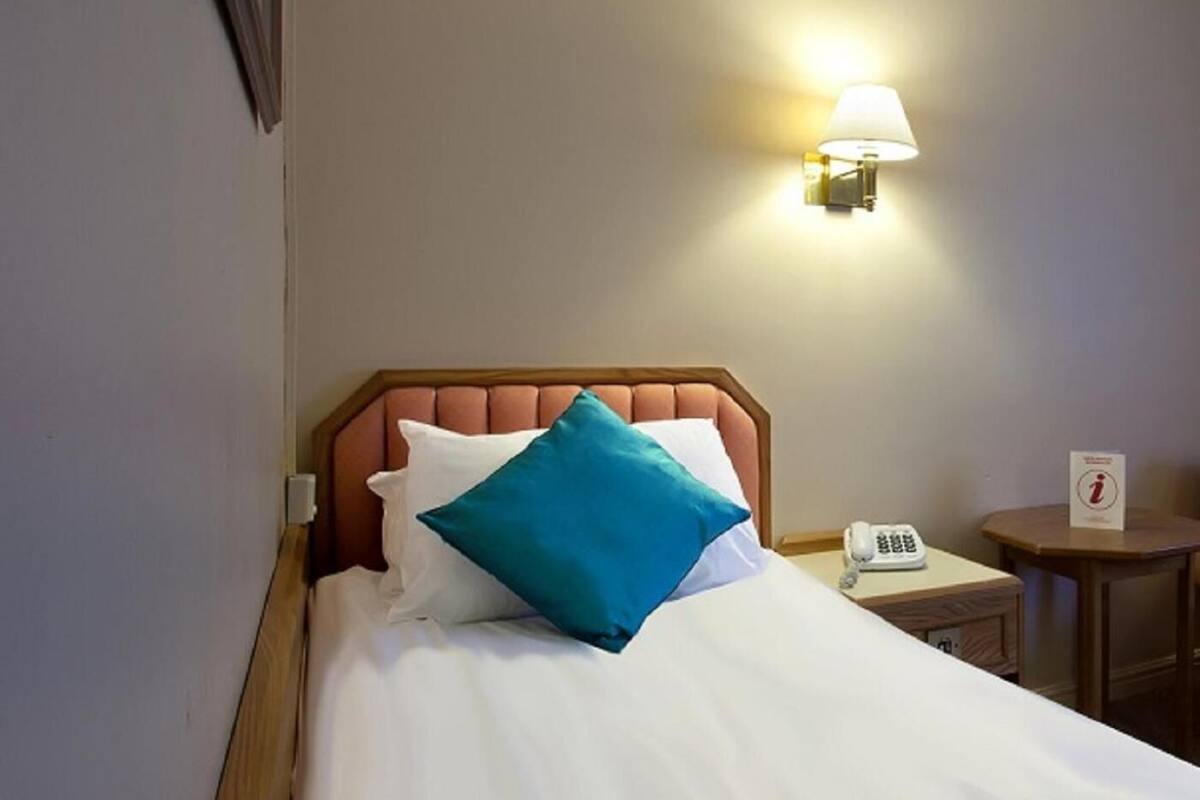 A hotel room with a bed, a bedside lamp, a phone, and a small table with a sign.