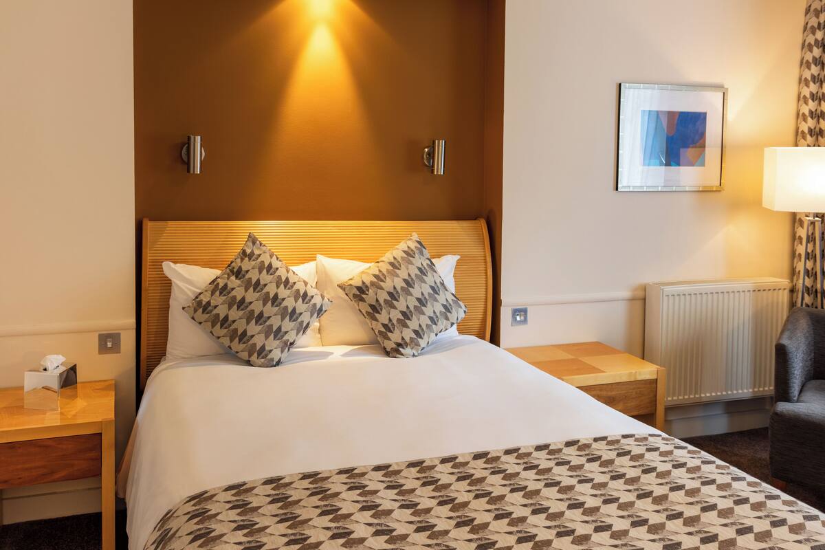 A hotel room with a large bed, two pillows, a bedside table with a lamp, a chair, and a framed picture on the wall.