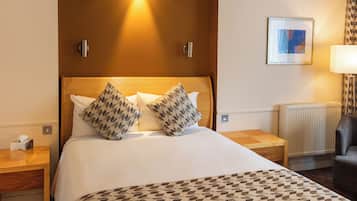A hotel room with a large bed, two pillows, a bedside table with a lamp, a chair, and a framed picture on the wall.