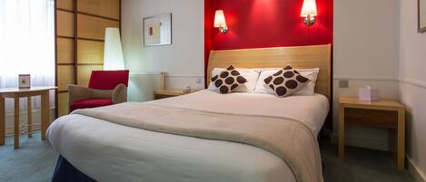 A hotel room with a large bed, two bedside lamps, a red accent wall, a wooden headboard, and a desk with a chair.