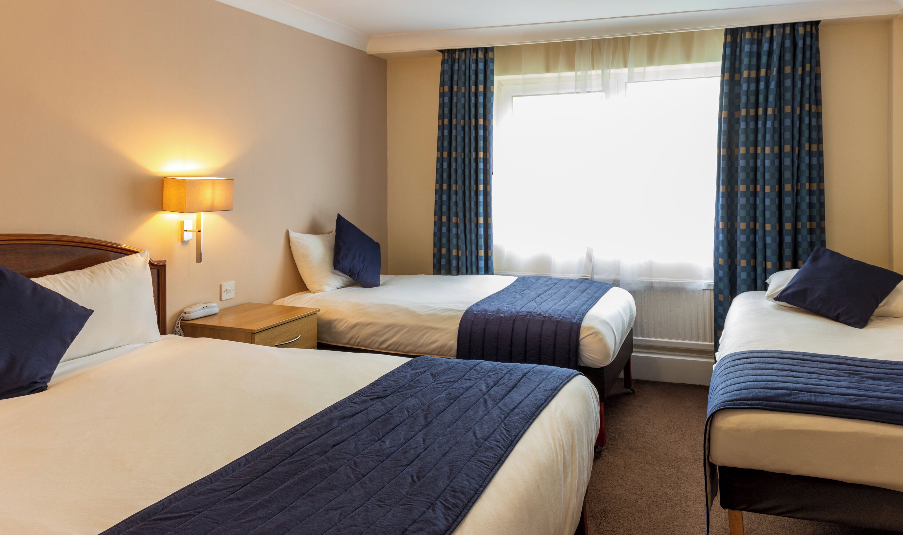 A hotel room with two beds, a nightstand, a lamp, and a window with curtains.