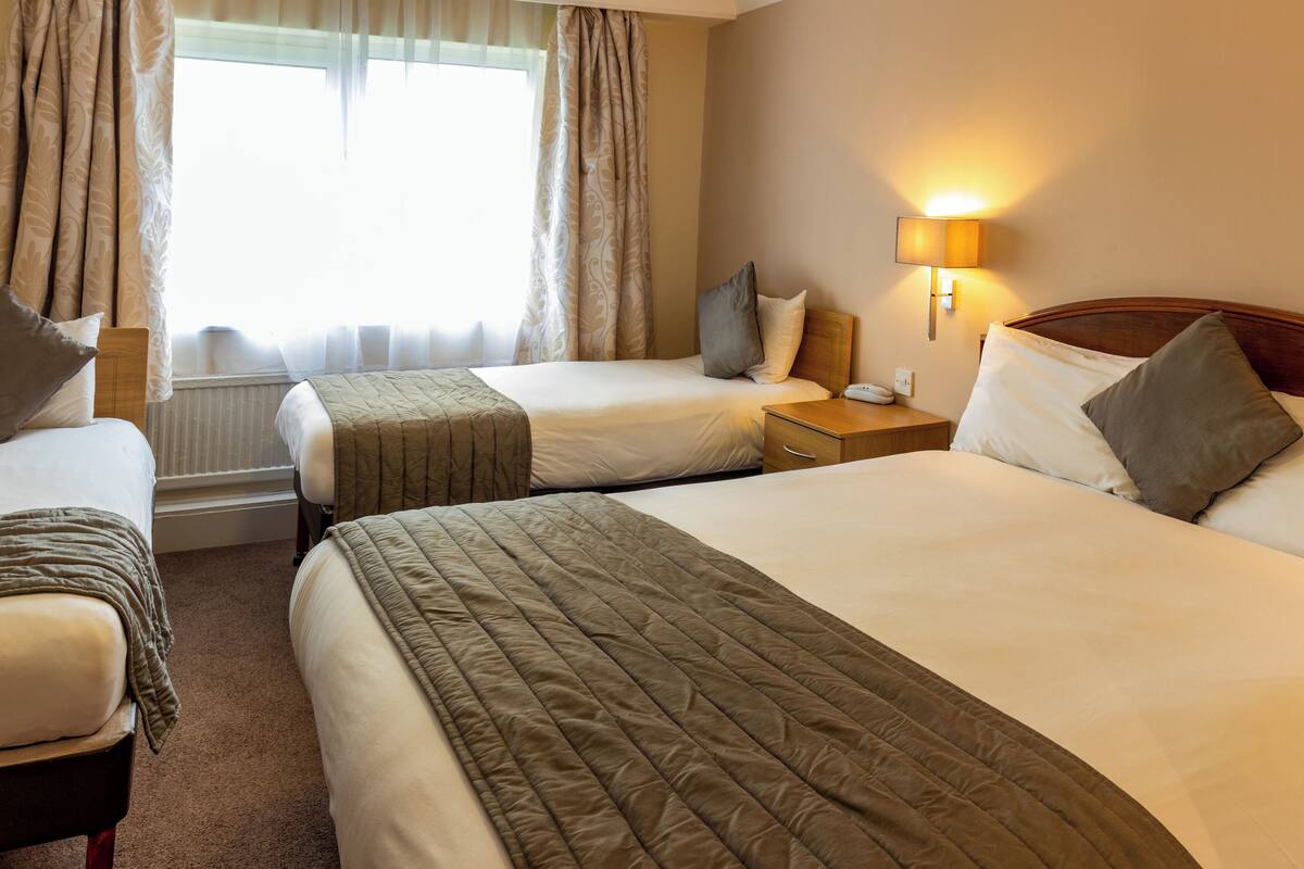 A hotel room with two beds, a bedside table with a lamp, and a window with curtains.