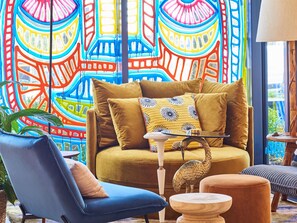 Una sala de estar acogedora con un mural abstracto llamativo, un sofá cómodo con cojines, un sillón azul y una mesa de centro de madera.