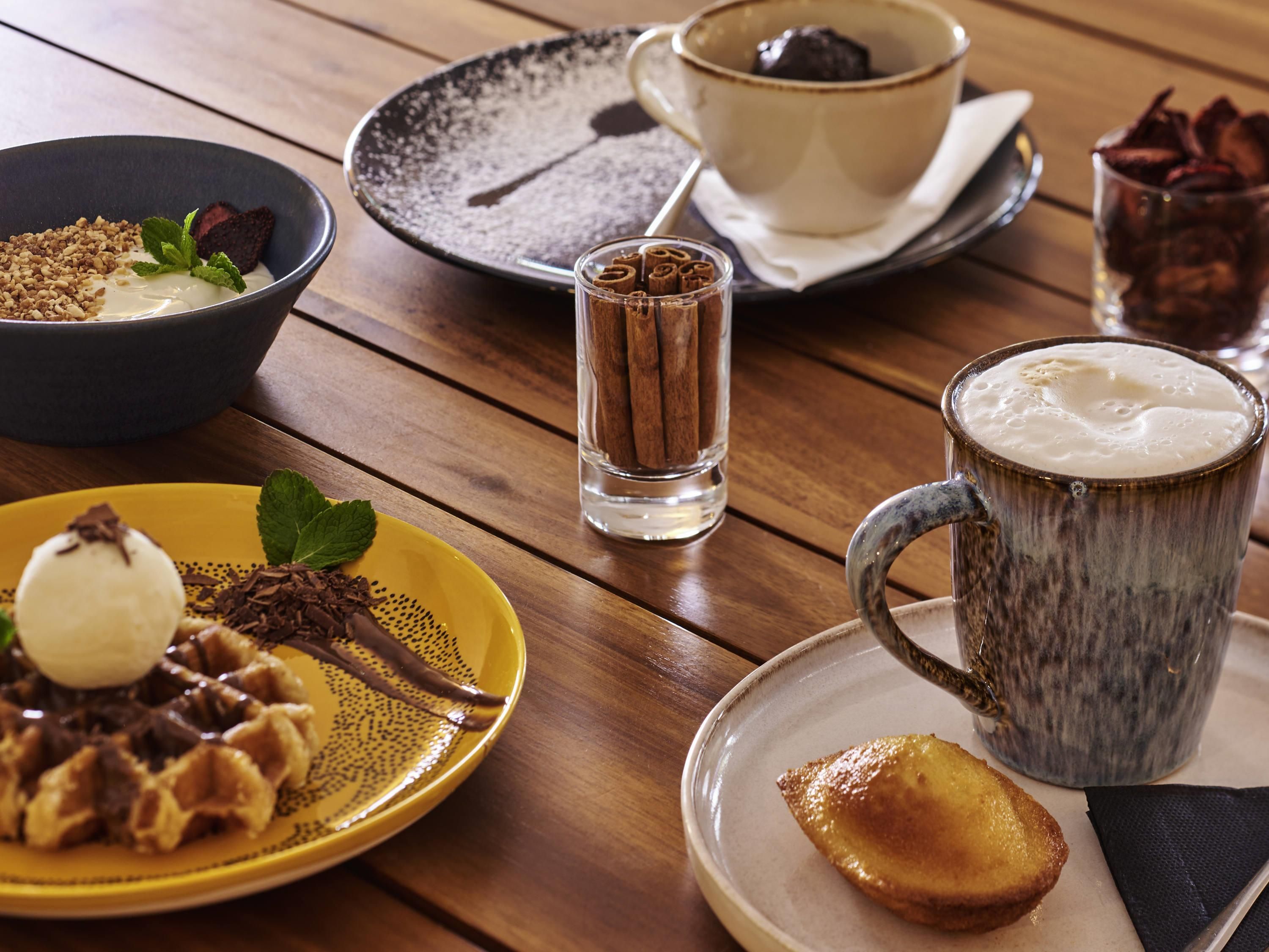 A wooden table with a variety of food items including a bowl, a plate with waffles, a mug, and a cup.