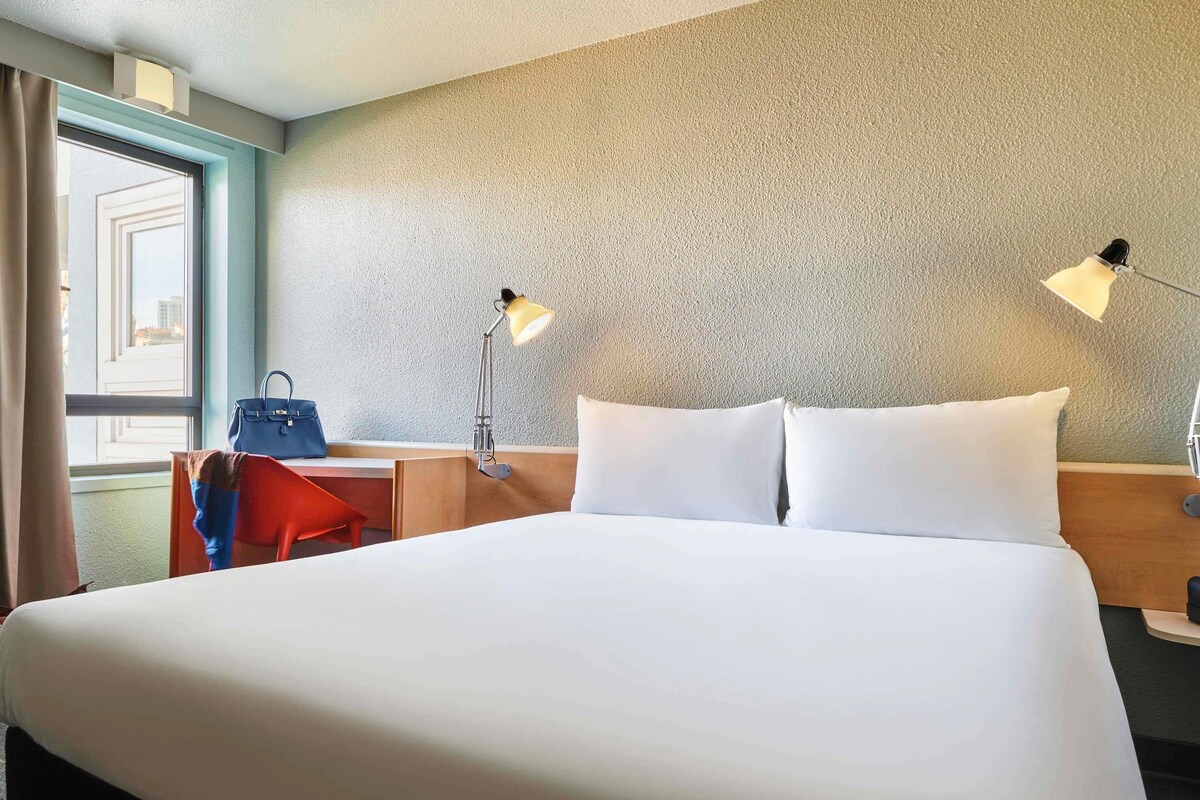 A hotel room with a large bed, two bedside lamps, a desk with a chair, a handbag, and a window with curtains.