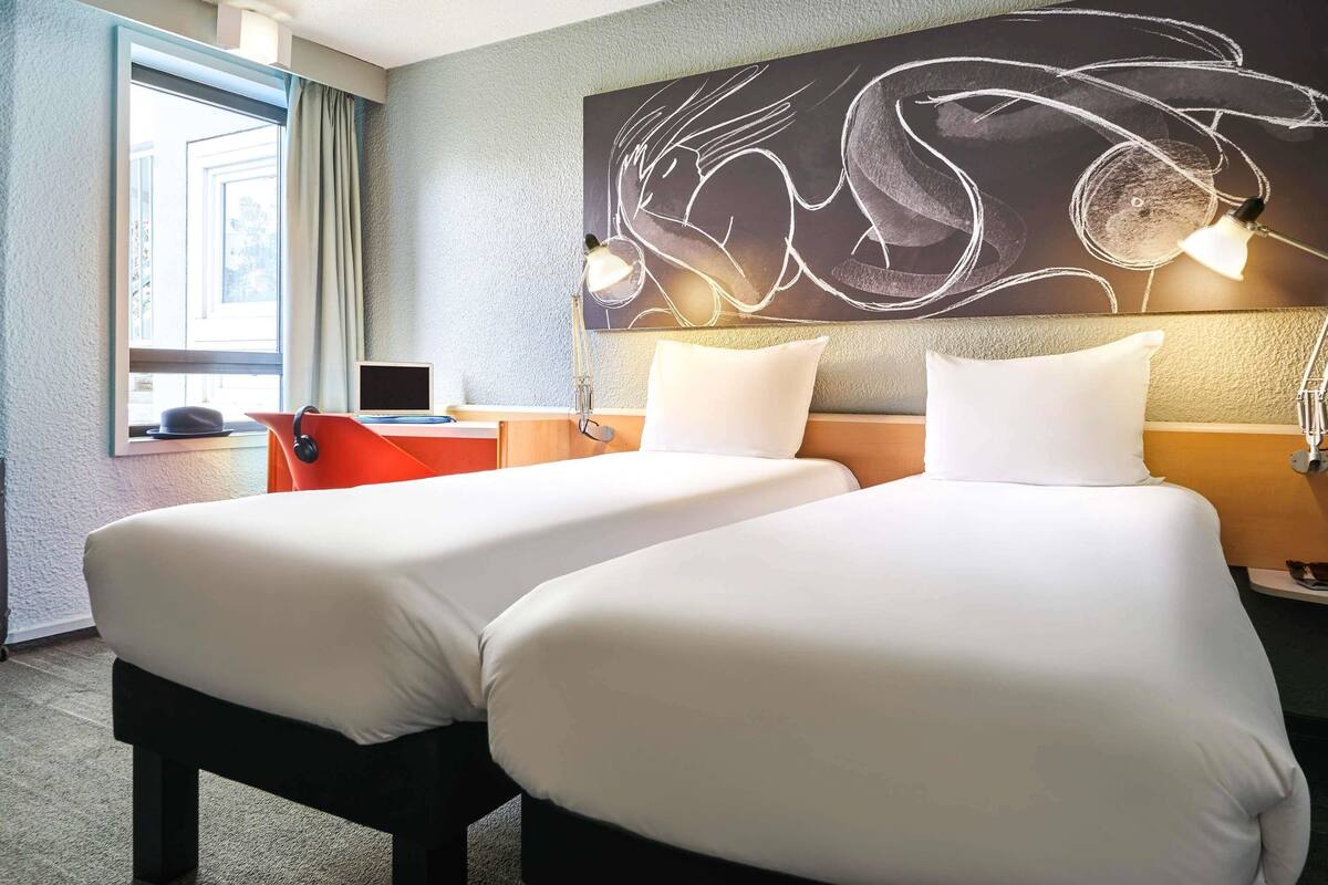 A hotel room with two beds, a desk with a laptop, a large abstract wall art, and a window with curtains.