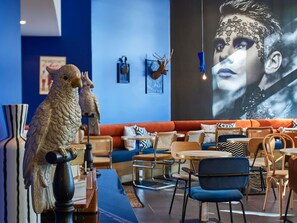 A modern interior with a blue wall, a large screen displaying a person, and a seating area with a sofa and chairs. - ibis Paris La Villette Cité des Sciences 19ème (Paris)