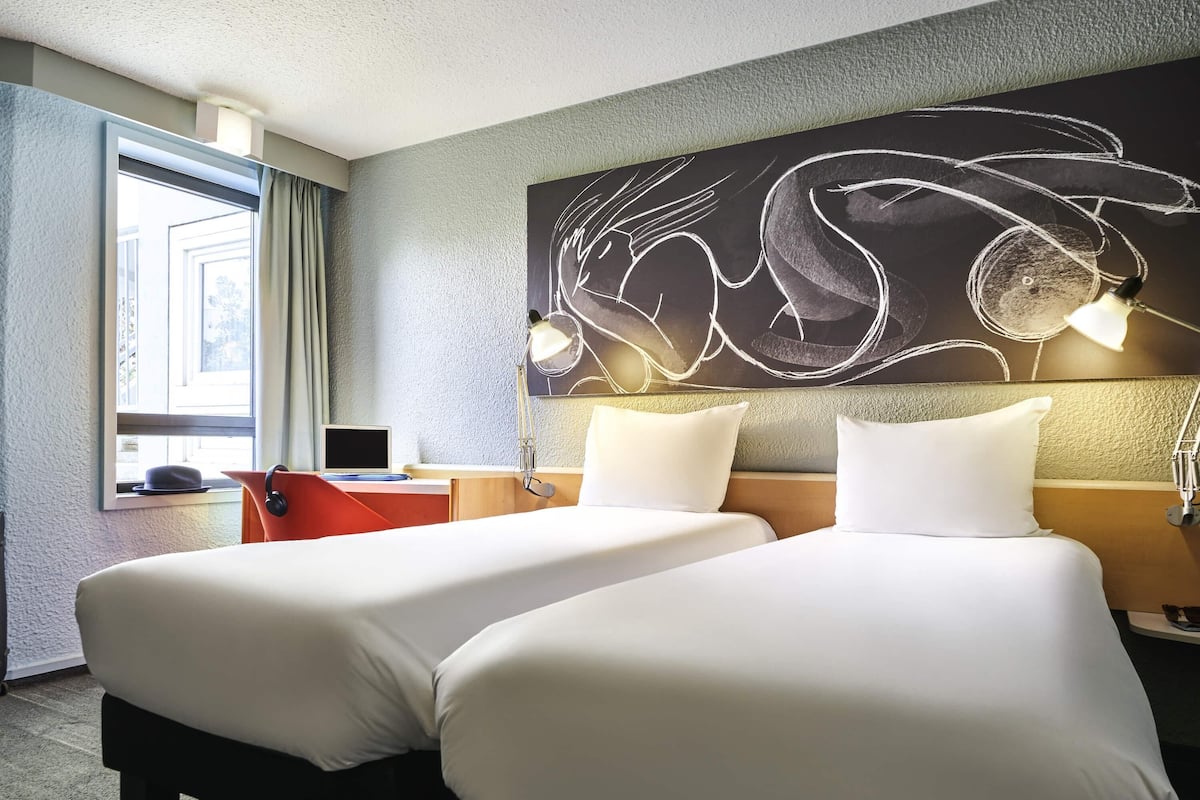 A hotel room with two beds, a desk, a laptop, a window with curtains, and a wall art piece.