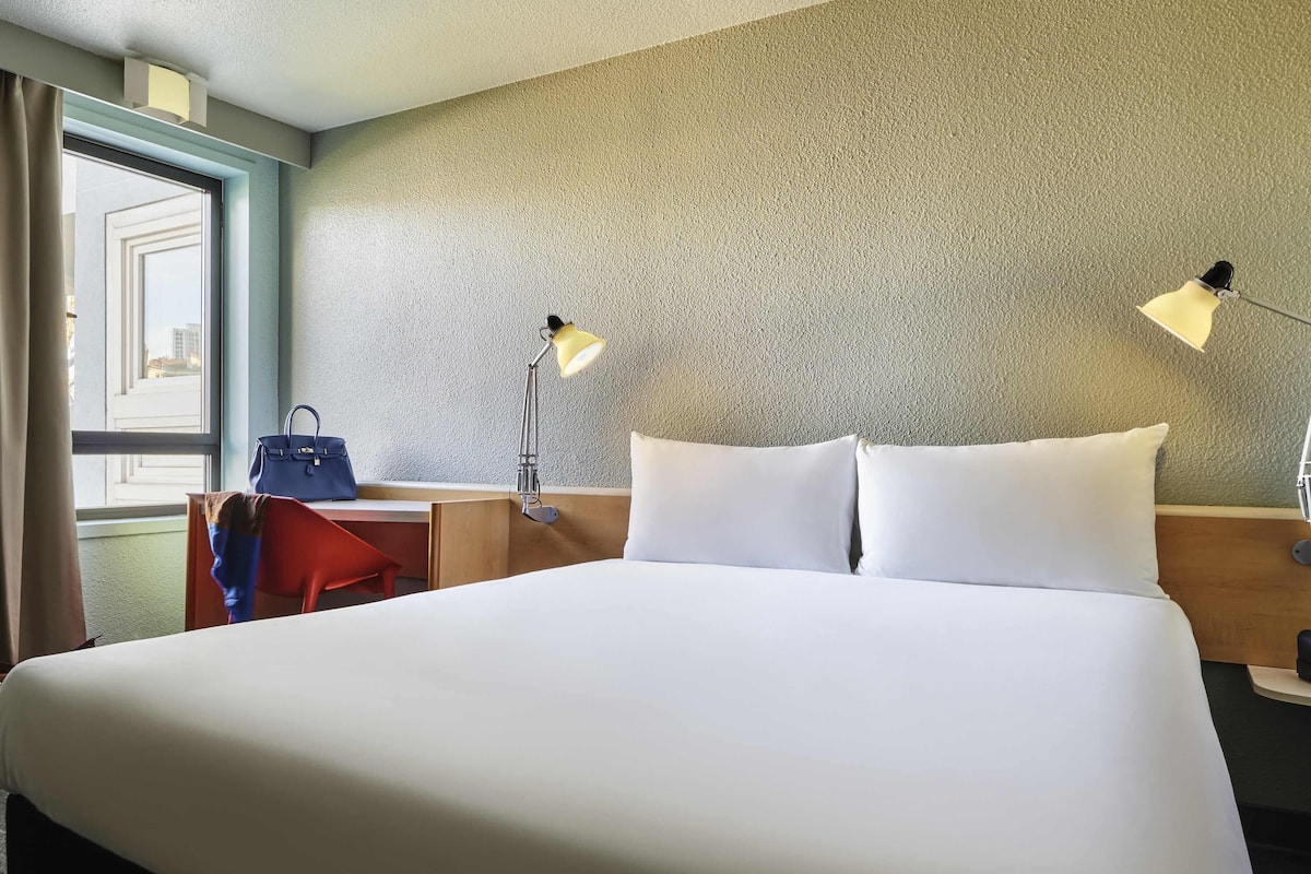 A hotel room with a bed, two bedside lamps, a desk with a chair, a window with curtains, and a view of buildings outside.