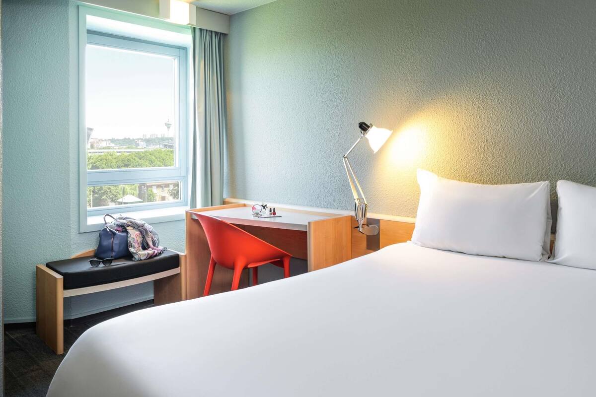 A hotel room with a bed, a desk, a chair, a lamp, a window, and a view of the city.