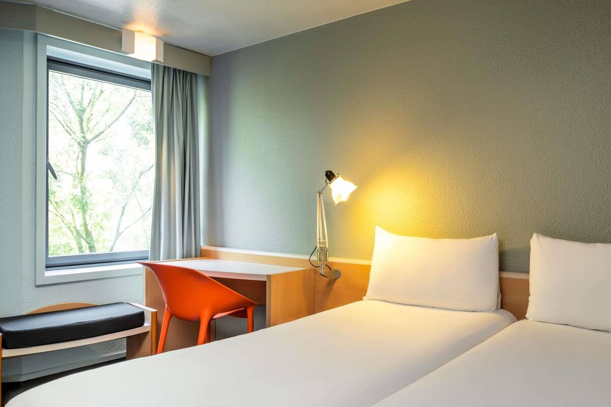 A hotel room with a bed, desk, chair, lamp, and a window with curtains.