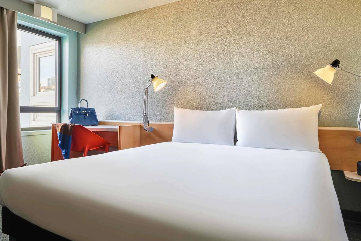 A hotel room with a large bed, two bedside lamps, a desk with a chair, a handbag, and a window with curtains.