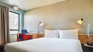 A hotel room with a large bed, two bedside lamps, a desk with a chair, a handbag, and a window with curtains. - ibis Paris La Villette Cité des Sciences 19ème (Paris)