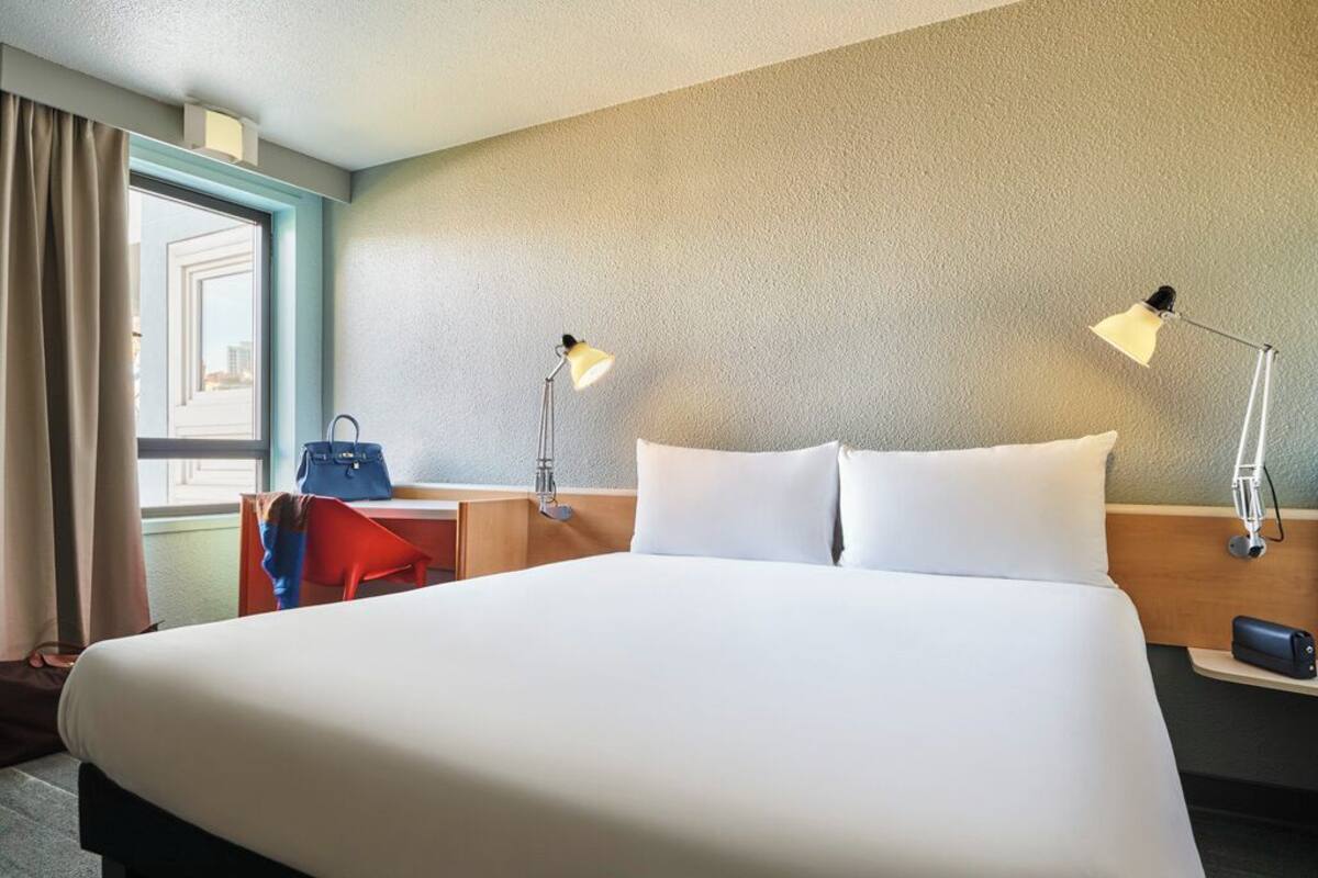 A hotel room with a large bed, two bedside lamps, a desk with a chair, a handbag, and a window with curtains.