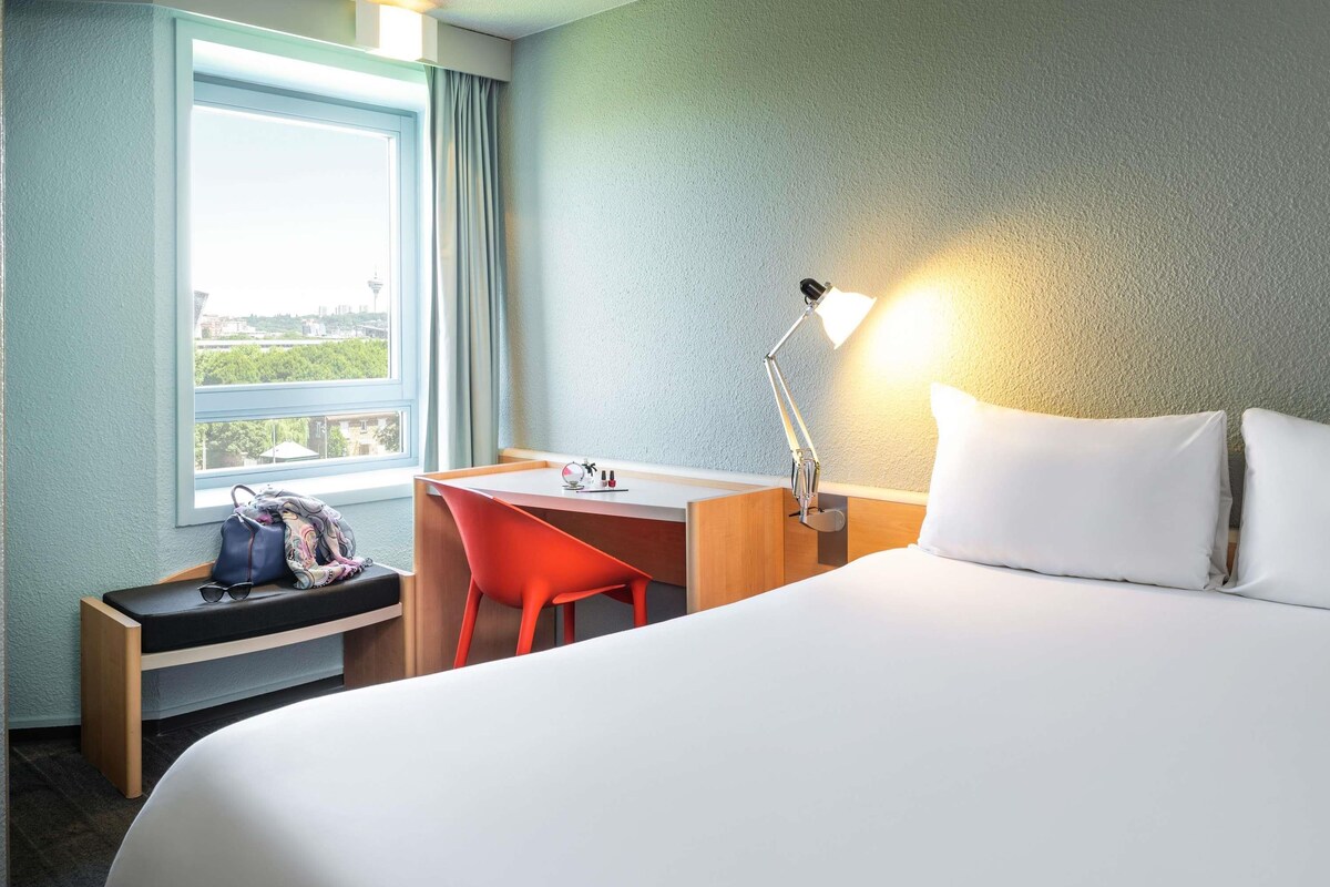 A hotel room with a bed, a desk, a chair, a lamp, a window, and a view of the city.