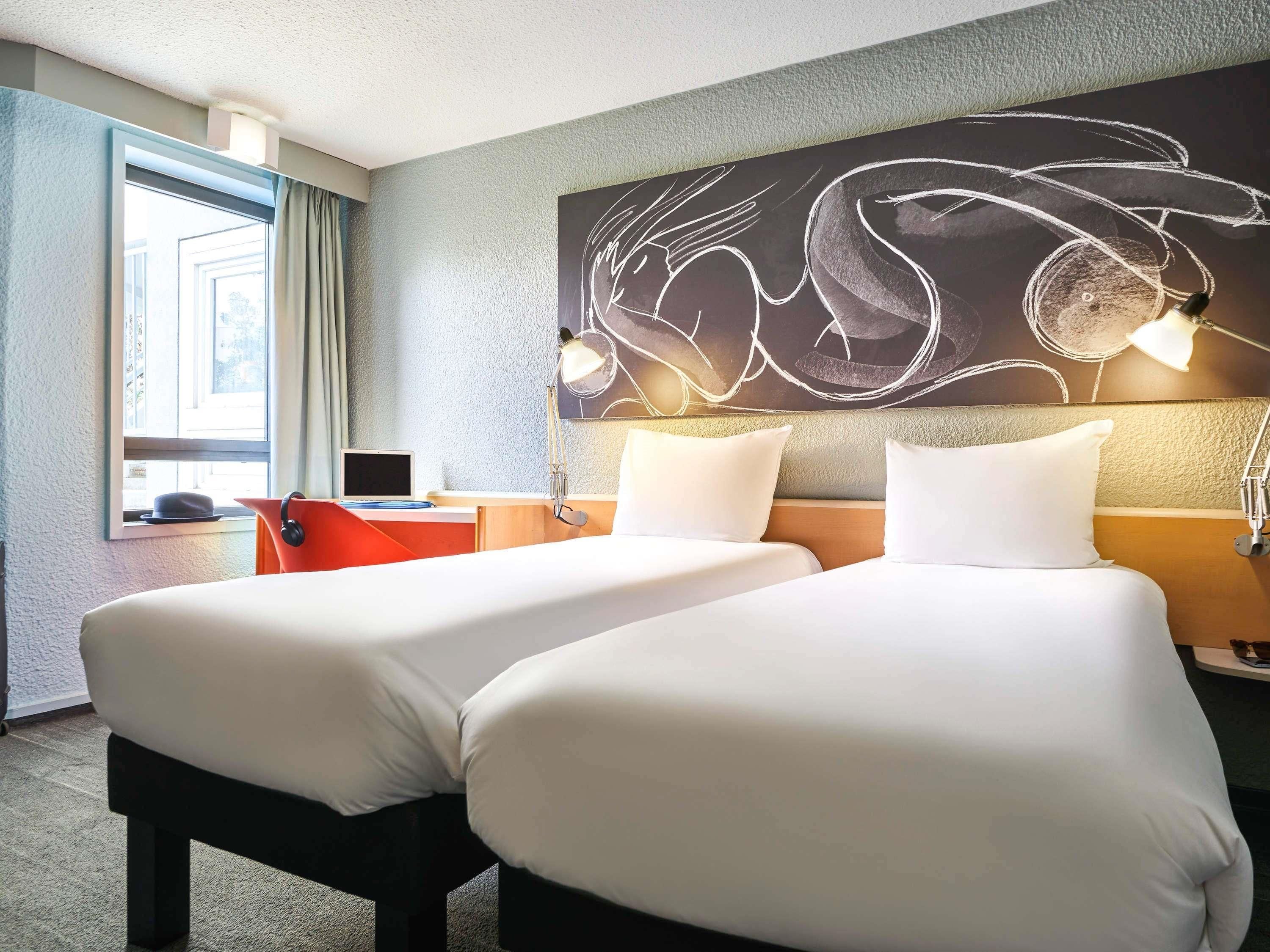 A hotel room with two beds, a desk with a laptop, a large abstract wall art, and a window with curtains.