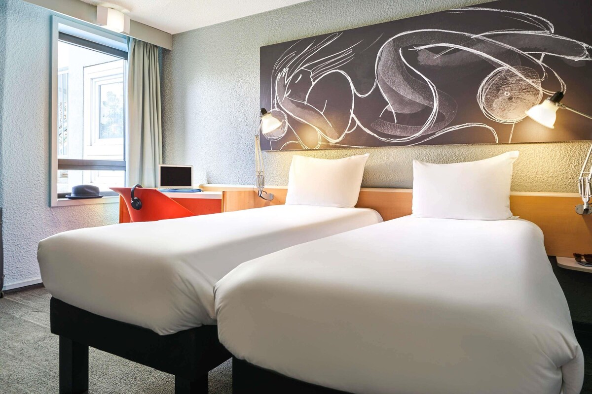 A hotel room with two beds, a desk with a laptop, a large abstract wall art, and a window with curtains.