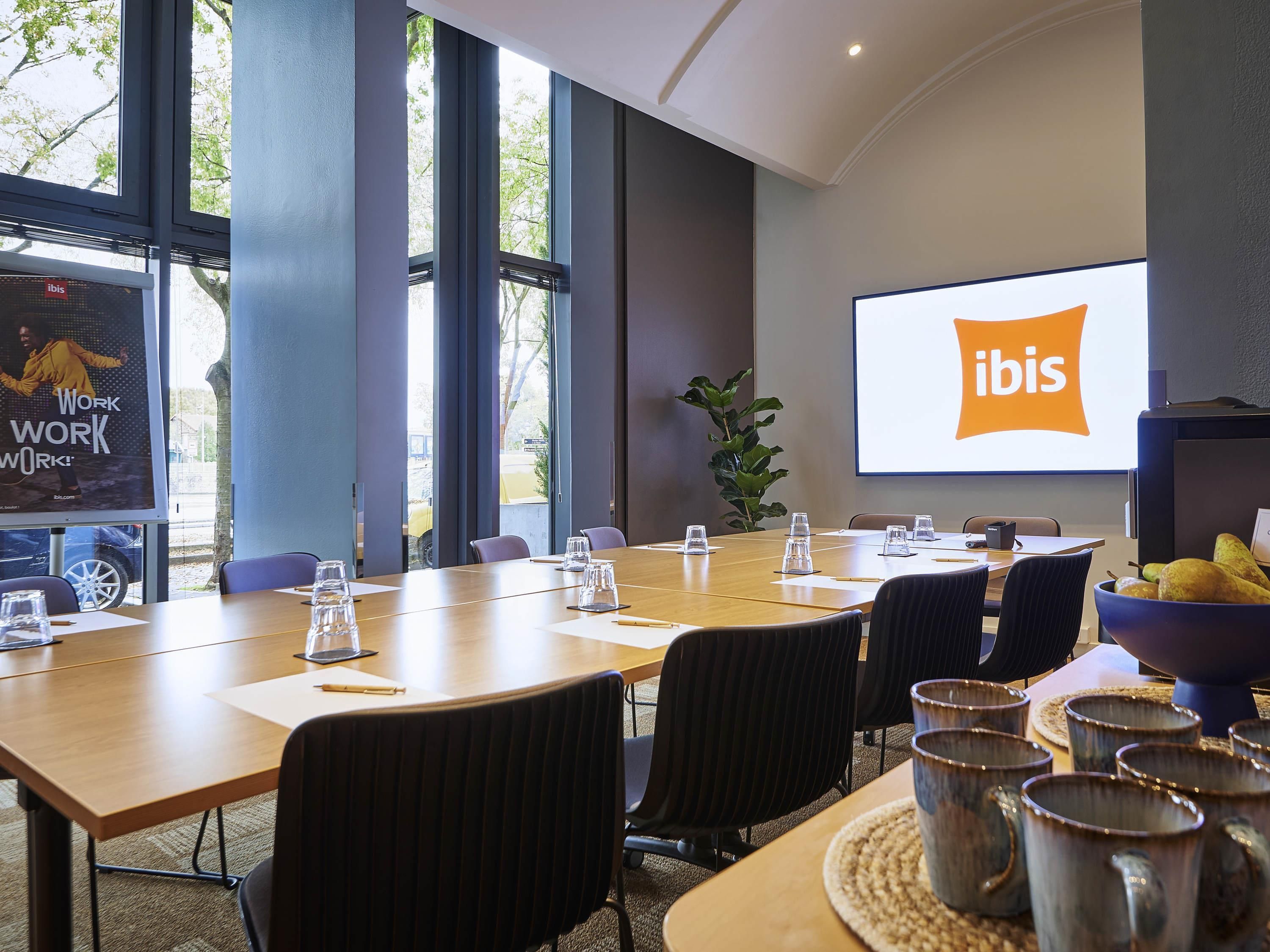 A conference room with a large screen displaying the ibis logo, a long table with chairs, and a plant.