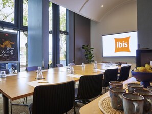 Un salón de conferencias con una pantalla grande mostrando el logotipo de ibis, una larga mesa con sillas y una planta.