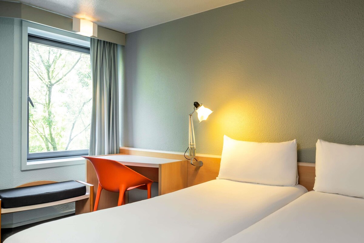 A hotel room with a bed, desk, chair, lamp, and a window with curtains.