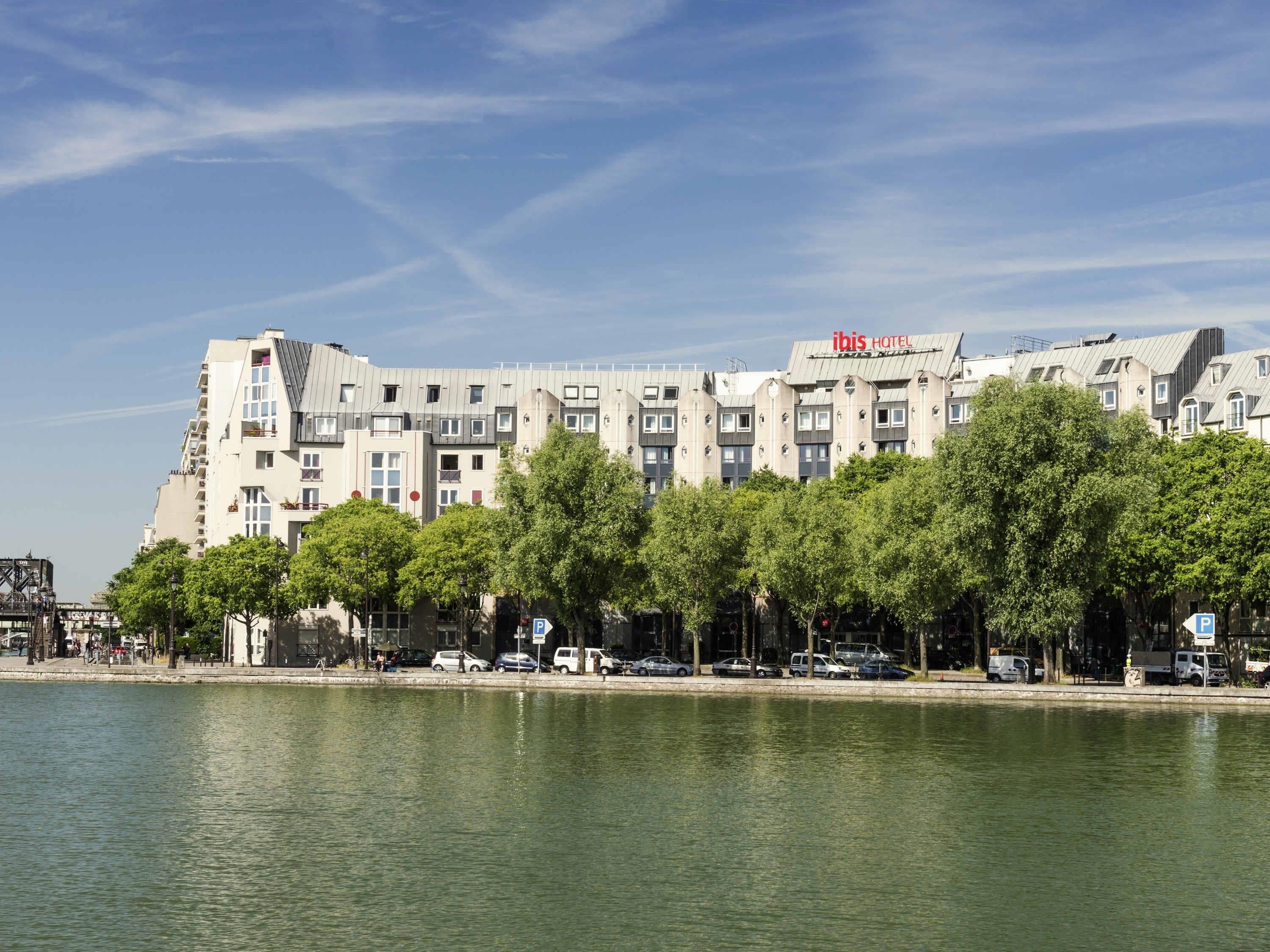Un hotel de varios pisos, con una señal visible que dice 'ibis HOTEL' y un cuerpo de agua al frente.