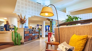A hotel lobby with a reception desk, a wall of suitcases, a cactus decoration, a lamp, and a sofa with a yellow pillow. - ibis Paris La Villette Cité des Sciences 19ème (Paris)