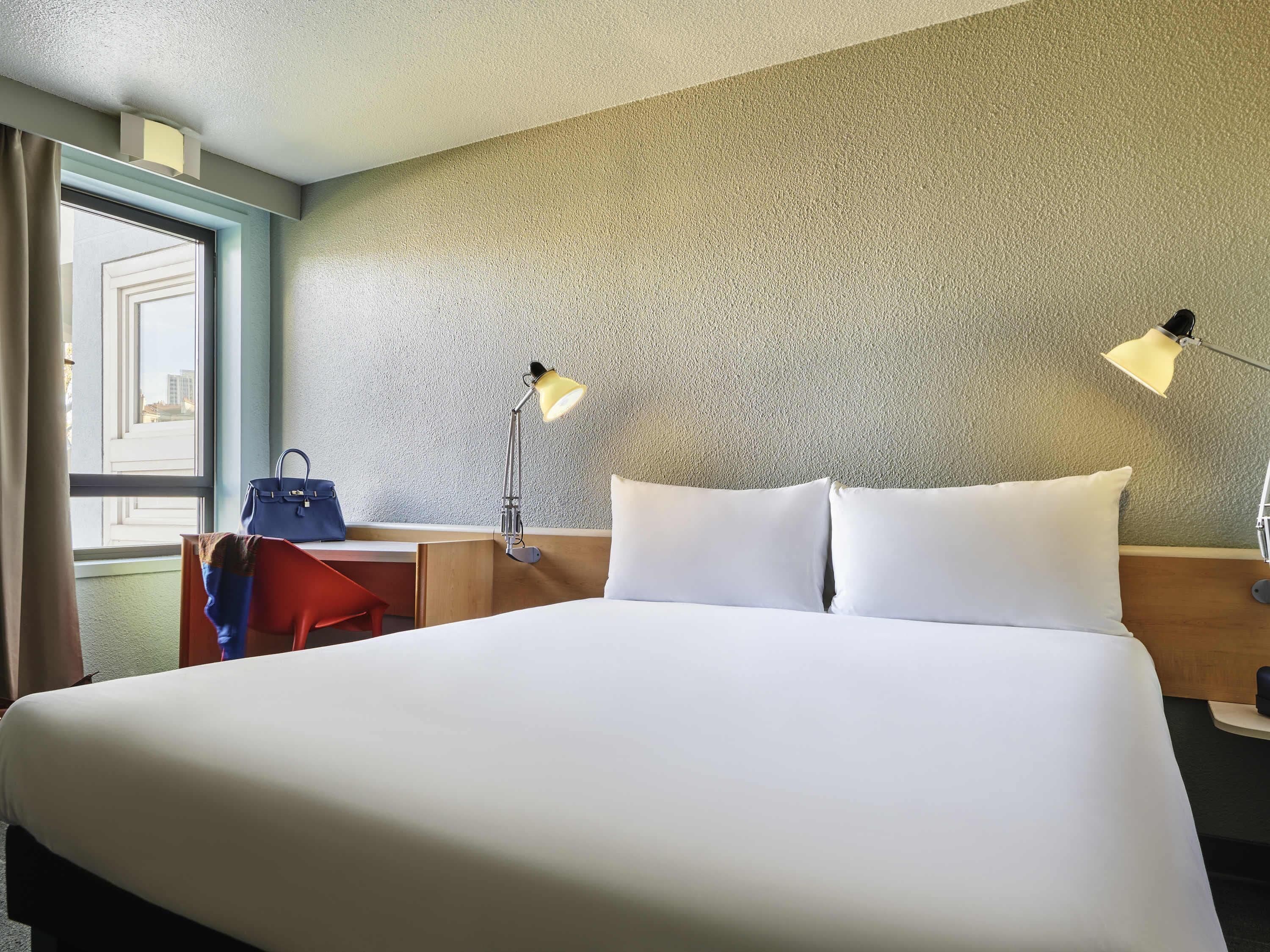 Ein Hotelzimmer mit einem Bett, zwei Nachttischlampen, einem Schreibtisch mit Stuhl, einem Fenster mit Vorhängen und Blick auf Gebäude.