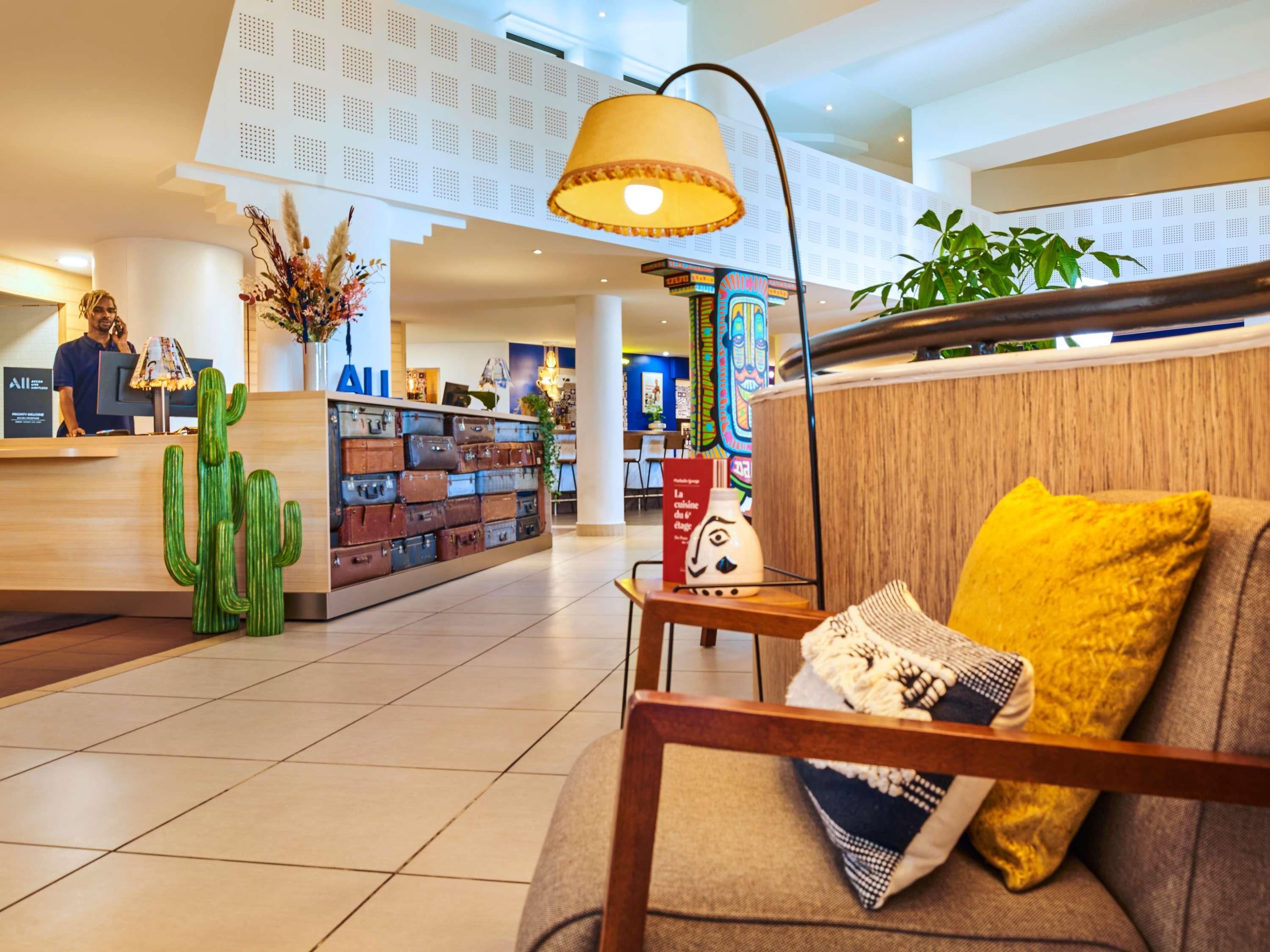 A hotel lobby with a reception desk, a wall of suitcases, a cactus decoration, a lamp, and a sofa with a yellow pillow.