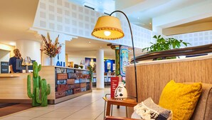 A hotel lobby with a reception desk, a wall of suitcases, a cactus decoration, a lamp, and a sofa with a yellow pillow. - ibis Paris La Villette Cité des Sciences 19ème (Paris)