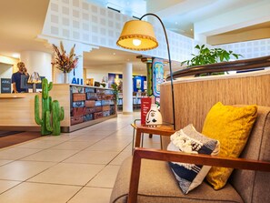 Un vestíbulo de hotel con una mesa de recepción, una pared de maletas, una decoración de cactus, una lámpara y un sofá con un cojín amarillo.