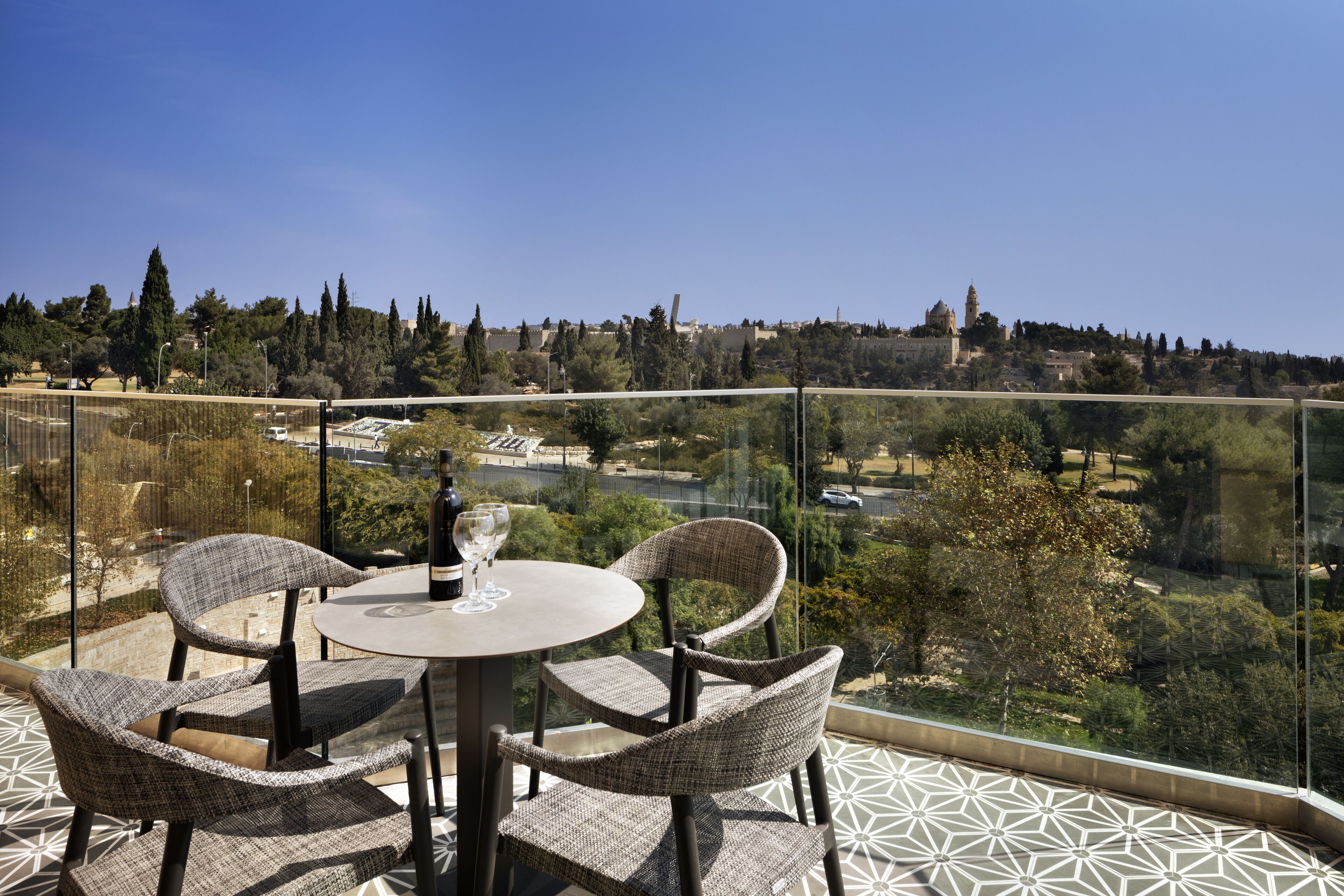 A balcony with a round table and wicker chairs, overlooking a scenic view of a city and hills.