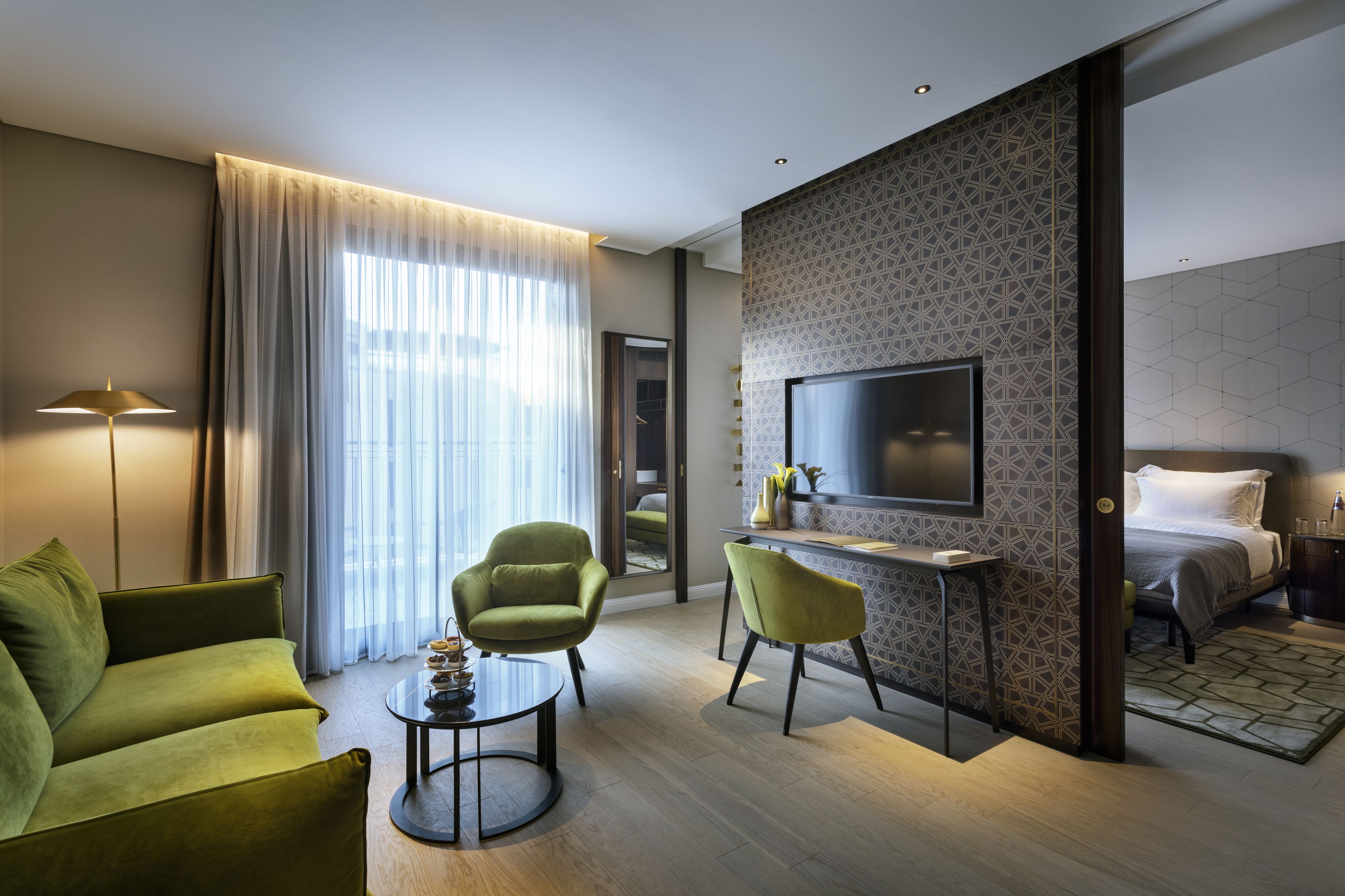 A modern hotel room with a sofa, two chairs, a desk, a TV, and a bed.