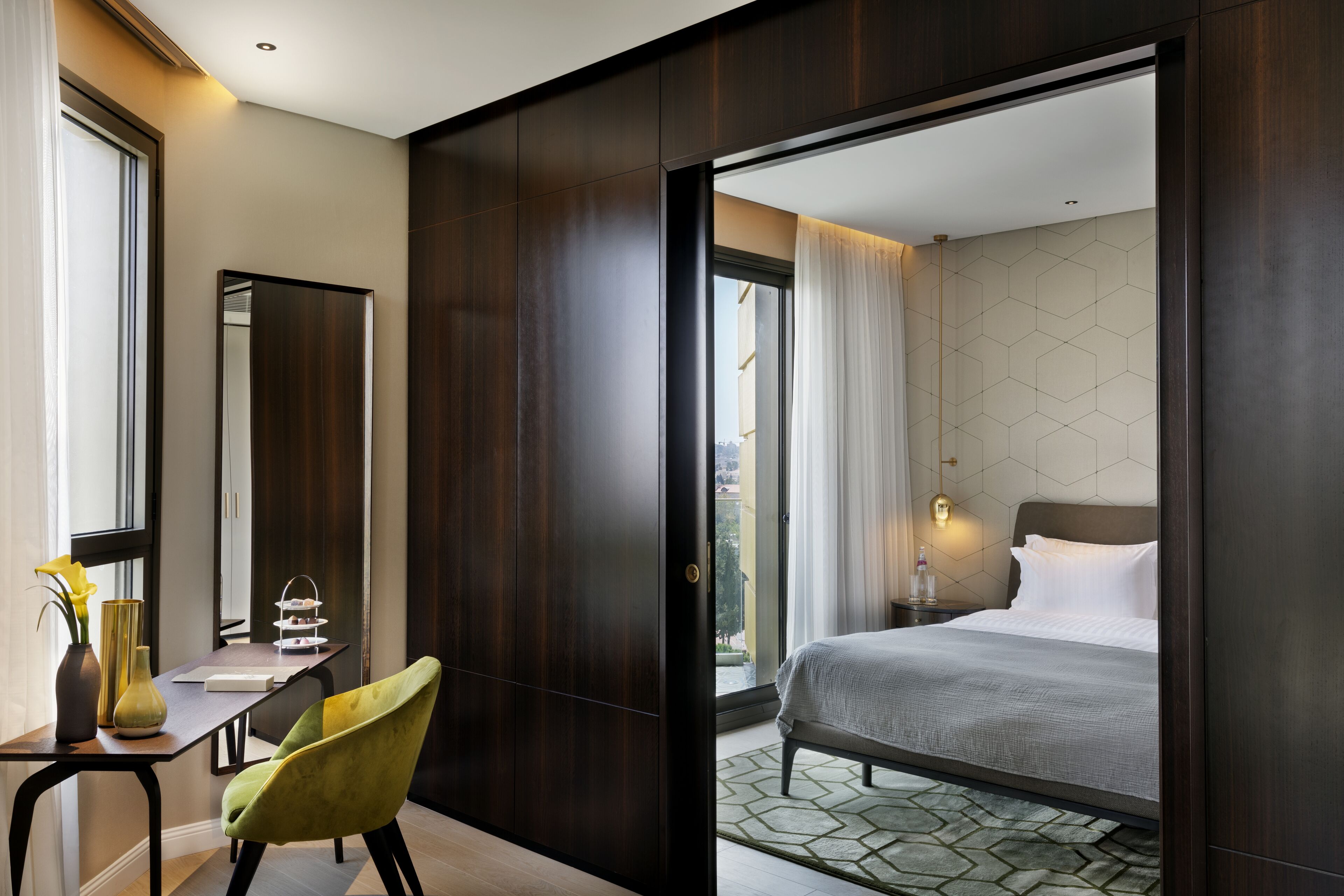 A modern hotel room with a large bed, a desk with a chair, a mirror, and a view of the city through a glass door.