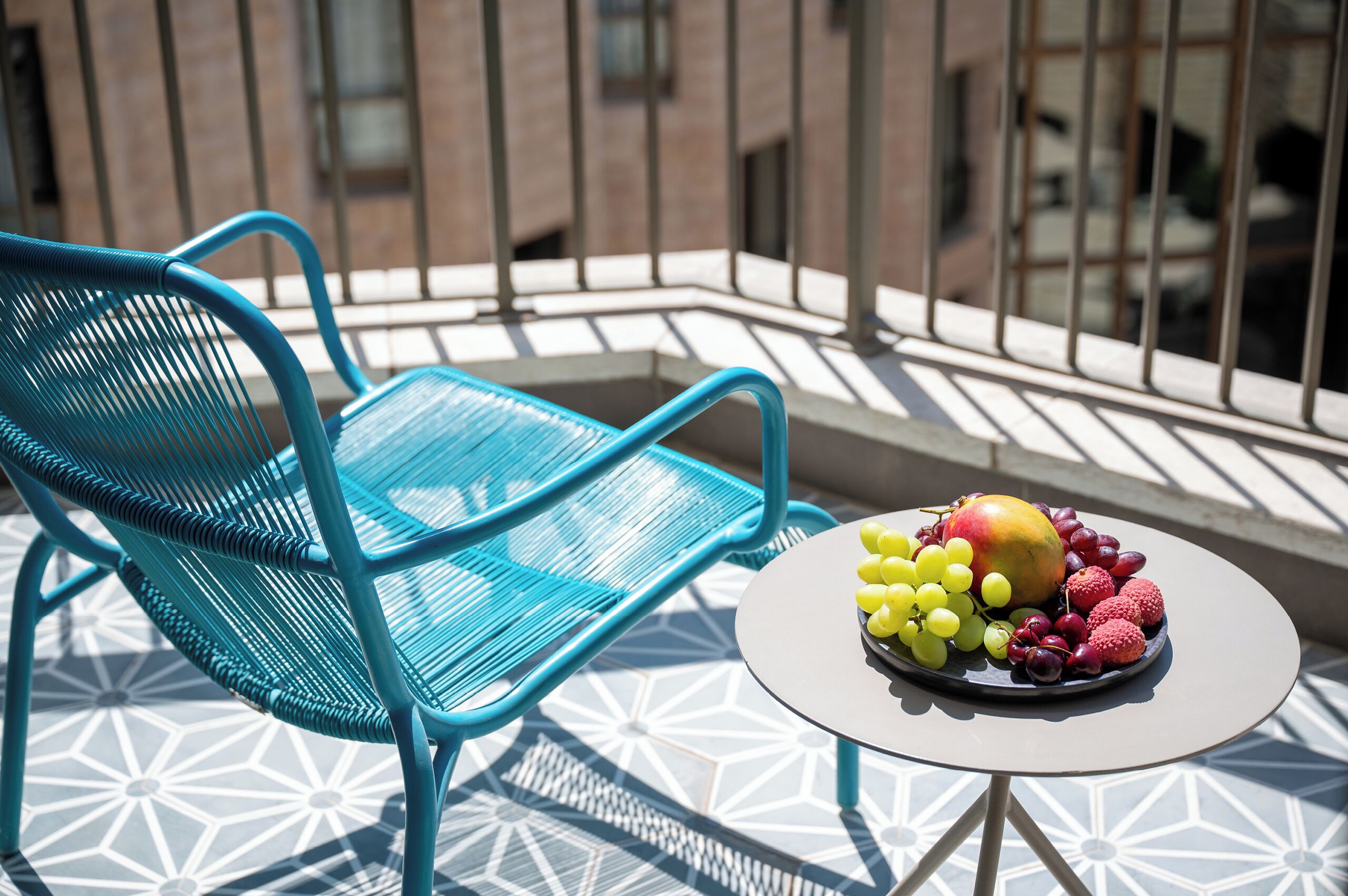 Une chaise en métal bleue et une petite table ronde sur laquelle se trouve un panier de fruits, le tout sur un balcon doté d’un sol à motifs et de rampes en métal.