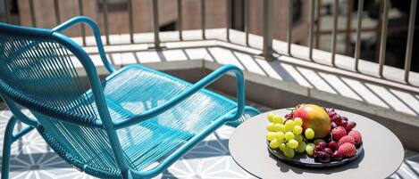 Une chaise en métal bleue et une petite table ronde sur laquelle se trouve un panier de fruits, le tout sur un balcon doté d’un sol à motifs et de rampes en métal.