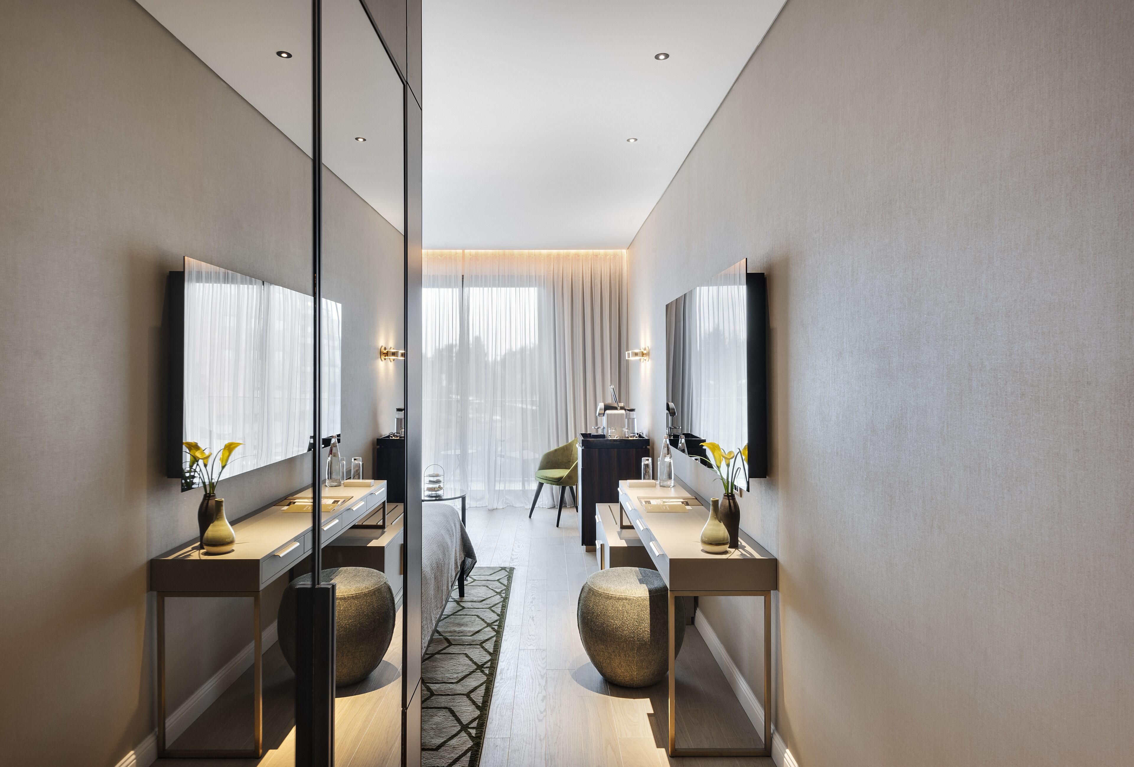 Une chambre d’hôtel moderne dotée d’un grand miroir, d’une coiffeuse avec un lavabo et d’un lit agrémenté d’un tapis à motifs géométriques.