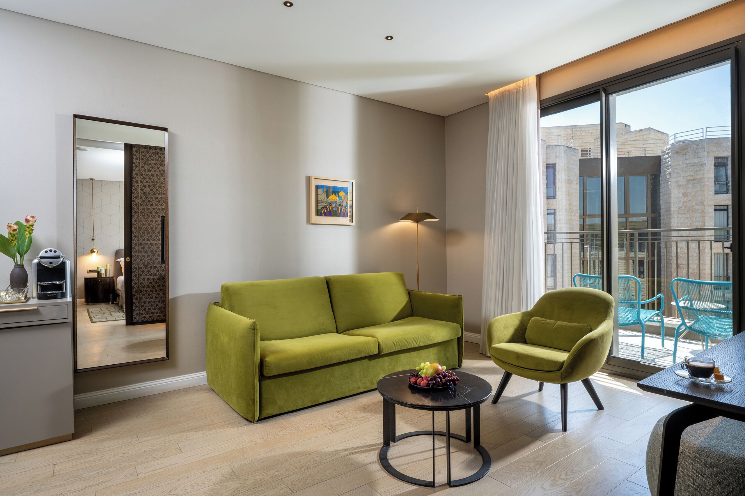 Una habitación de hotel moderna con un sofá verde, una pequeña mesa redonda con fruta, un sillón verde, un gran ventanal con balcón y un espejo con diseño geométrico.