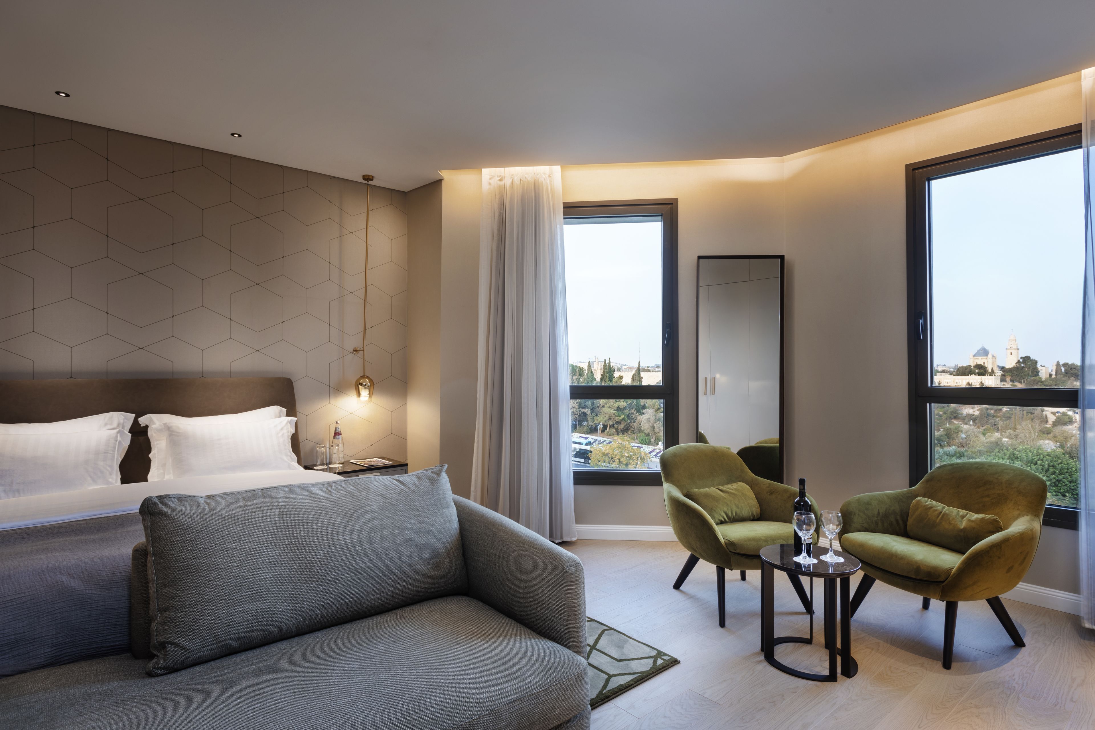 A modern hotel room with a large bed, a sofa, two armchairs, a small table with a bottle and glass, and large windows offering a city view.