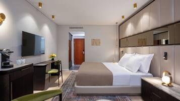 A modern hotel room with a large bed, a desk with a flat-screen TV, a coffee machine, and a small seating area with a chair and a table.