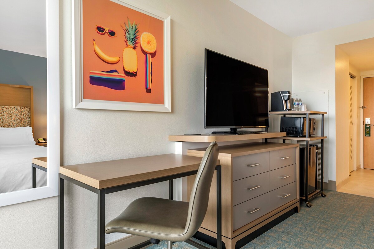 A hotel room with a bed, a desk with a chair, a flat-screen TV, a coffee machine, and a framed artwork on the wall.