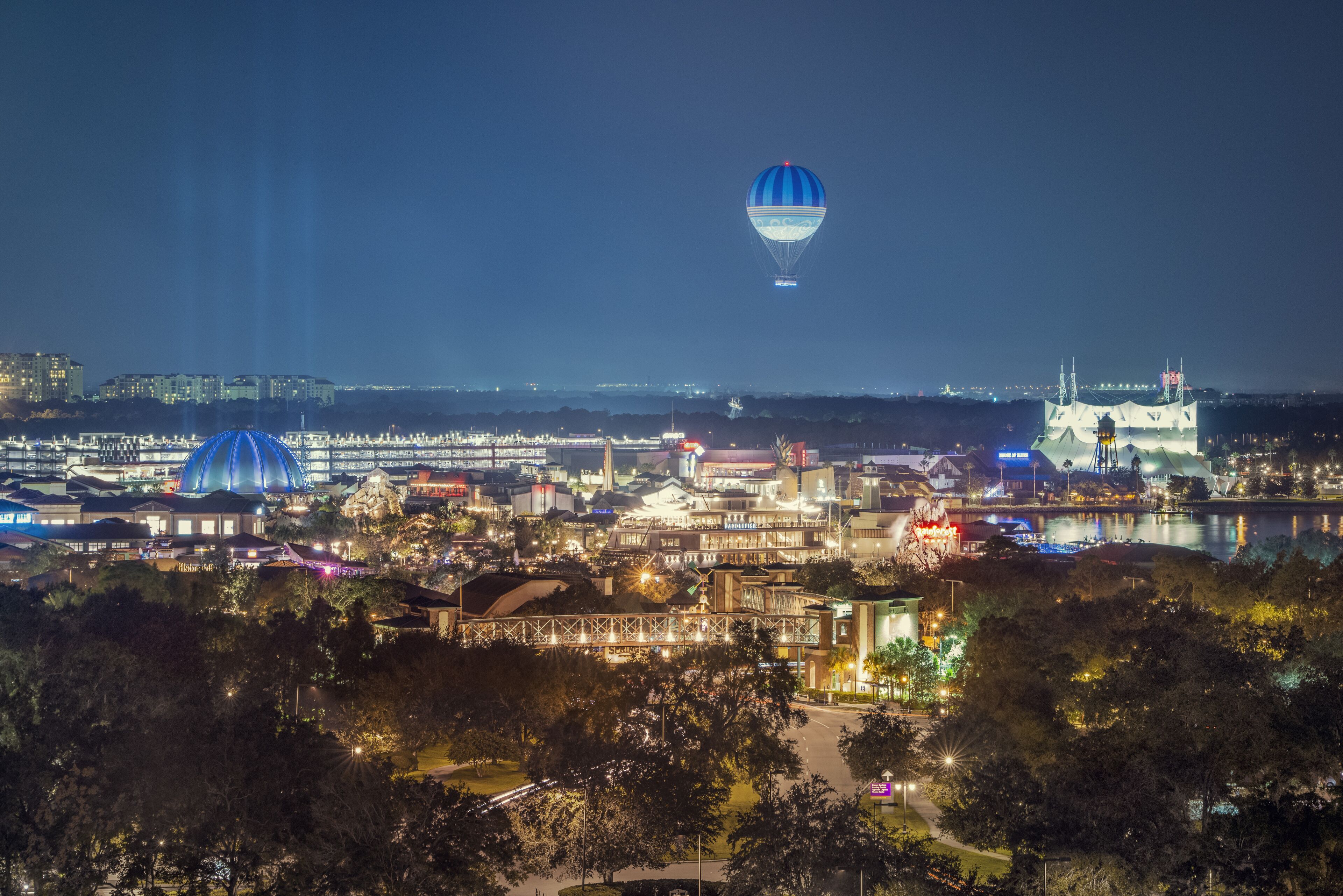 A nighttime cityscape with illuminated buildings, a prominent dome structure, and a hot air balloon in the sky.